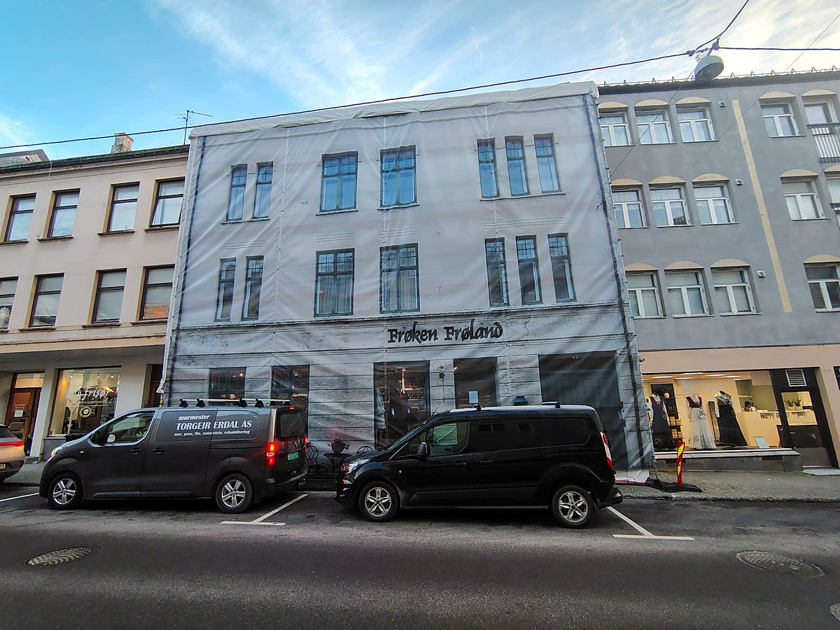 Bygg i sentrum kledt i stillas med trykt fasade og parkerte servicebiler