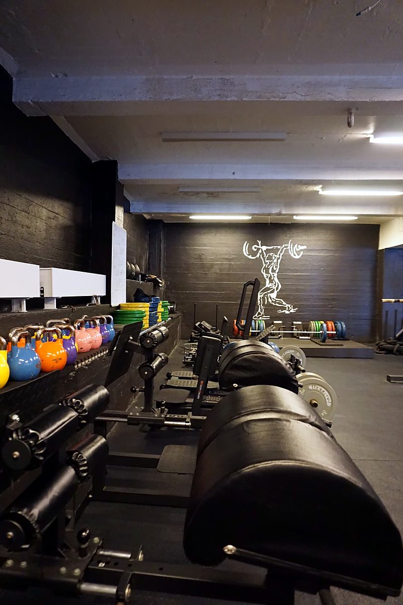 Treningssal med apparater, fargerike kettlebells langs veggen og en hvit vektløfterfigur malt på endeveggen