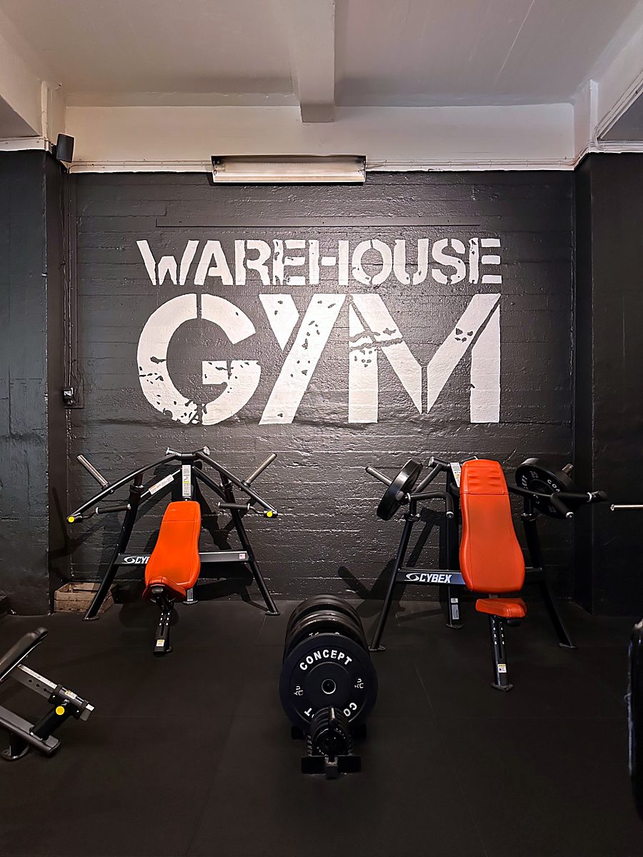To treningsapparater med oransje seter foran en sort vegg med teksten Warehouse Gym
