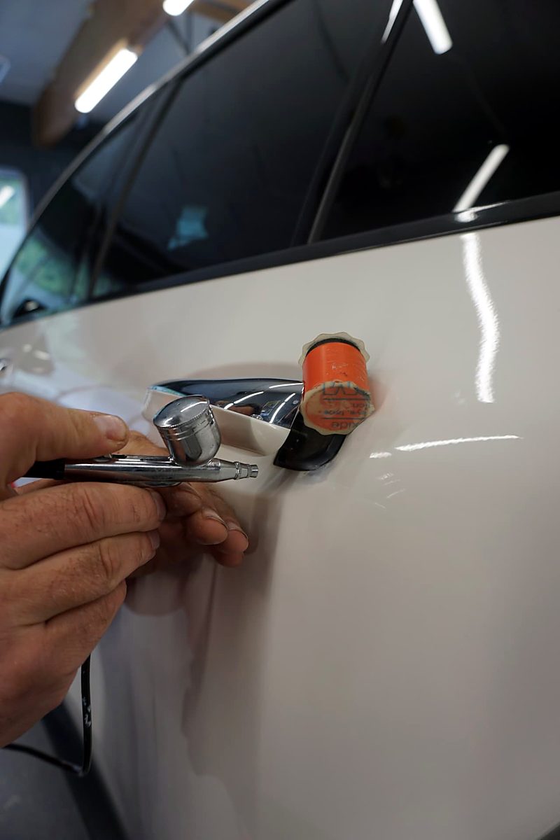 Hender som lakerer et hvitt bilpanel med airbrush, med oransje maskering ved dørvant